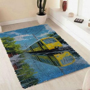 Puede incluir: Una alfombra rectangular con un tranvía amarillo reflejado en el agua. La alfombra tiene un fondo azul con un cielo y árboles. El tranvía es el punto focal, con su reflejo creando una imagen simétrica.