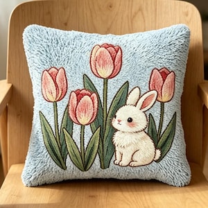 Puede incluir: Un cojín cuadrado con una superficie texturizada de color azul claro que presenta un diseño bordado de tres tulipanes rosas con tallos y hojas verdes junto a un pequeño conejo blanco con orejas y mejillas rosas sentado.