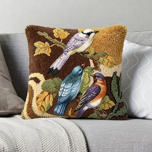 Puede incluir: Un cojín cuadrado con un diseño texturizado y copetudo de tres pájaros posados en ramas con hojas. Los pájaros están coloreados en tonos blanco, azul y naranja, sobre un fondo de hilo de color marrón y amarillo mostaza.
