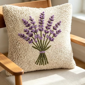 May include: A square throw cushion with a cream-coloured, textured, tufted surface featuring a central embroidered design of a lavender bouquet. The lavender flowers are purple with green stems and leaves, tied together with a small purple band.