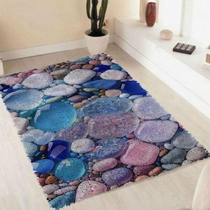 May include: A rectangular rug with a stone design. The rug features a pattern of various-sized stones in shades of blue, gray, and pink. The stones are arranged closely together, creating a textured appearance. The rug has a fringed edge.