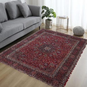 Könnte beinhalten: Ein rechteckiger, roter Perserteppich mit einem komplizierten Blumenmuster und einem dunklen Rand. Der Teppich liegt auf einem hellen Holzboden in einem Wohnzimmer. Das Design des Teppichs umfasst ein zentrales Medaillon und detaillierte Eckmotive.