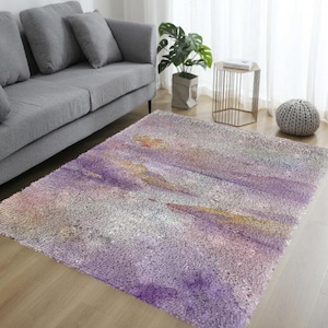 May include: A rectangular area rug with a soft, textured surface. The rug features a watercolor-style design with shades of purple, gray, and gold. The rug is placed on a hardwood floor in a living room setting.
