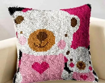 May include: Square decorative pillow with a textured, plush surface. The design features a cartoon bear with a brown face, pink cheeks, and a pink heart. The bear is set against a white and pink background.