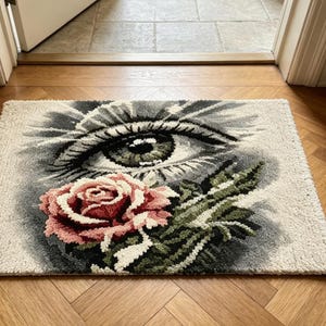 May include: A rectangular tufted rug featuring a detailed graphic design of a large, realistic human eye in shades of gray and black, positioned above a prominent, textured pink rose with green leaves, all set against a cream-colored background.