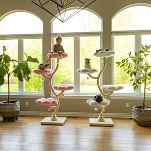 May include: Two tall cat trees with multiple levels shaped like flowers and clouds. One tree features pink, petal-shaped platforms, while the other has white, cloud-shaped platforms. Both structures have light-colored, curved vertical supports.