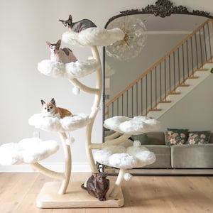Puede incluir: Un árbol para gatos blanco con múltiples plataformas con forma de nubes. El árbol para gatos tiene una base de madera marrón y está decorado con pompones blancos. Cuatro gatos están sentados en las plataformas y un quinto gato está sentado en la base del árbol.
