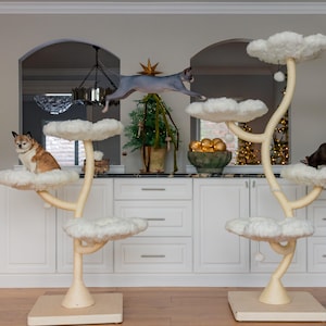 May include: Two cat trees with cloud-shaped platforms and cream-colored branches. One tree has a cat sitting on a platform, another has a cat jumping between platforms, and a third cat is on the other tree. The background includes a white cabinet and a countertop.