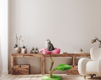 May include: A pink cat tree shaped like a flower with a gray cat sitting on the top platform. The cat tree has a green base and a brown wooden trunk. The cat tree is in front of a wooden shelf with a white wall behind it.