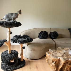 May include: A gray Sphynx cat perches atop a multi-level cat tree with faux fur platforms and a natural wood frame. The cat tree is in a living room with a beige sofa, black knot pillows, and a unique wooden coffee table.
