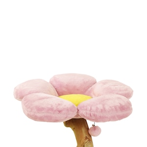 May include: A pink flower-shaped cat bed with a yellow center and a pink pom-pom hanging from the bottom. The bed is attached to a wooden post.