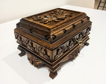 May include: A dark brown wooden box with intricate carvings and metal accents. The box has a hinged lid and a small keyhole. The box is sitting on a white surface.