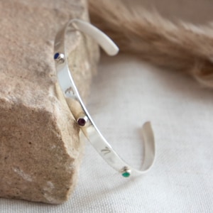 May include: A silver-toned cuff bracelet featuring three small, circular inset stones in blue, deep red, and green. The metal band has a smooth, polished finish and includes engraved letters positioned between the colourful stone accents.
