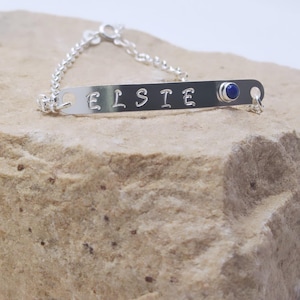 Può includere: Un braccialetto a catena d'argento con una targhetta personalizzata con il nome "ELSTE" e un piccolo accento di pietra preziosa blu.