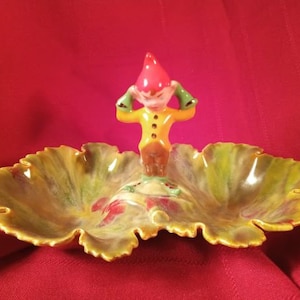 Puede incluir: Un plato de cerámica con forma de hoja con un esmalte verde, amarillo y marrón. Una pequeña figura de gnomo con un sombrero rojo y ropa amarilla y verde está de pie en el centro del plato.