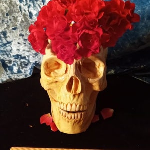 May include: A white ceramic skull with a red flower crown. The skull has a toothy grin and is decorated with a floral arrangement of red roses.