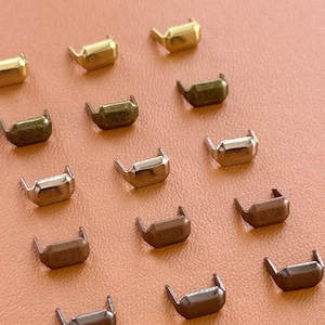 Puede incluir: Una colección de remaches metálicos en varios acabados, oro, bronce y plata, dispuestos sobre una superficie color melocotón. Estos pequeños sujetadores decorativos tienen un diseño curvo en forma de U, ideales para marroquinería y accesorios.