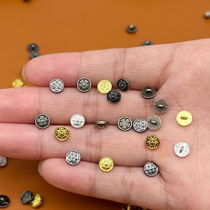 Puede incluir: Una colección de pequeños botones decorativos en varios acabados metálicos, incluyendo plata, oro y negro. Algunos botones presentan un diseño floral, mientras que otros son lisos. Los botones están dispersos sobre una superficie marrón.