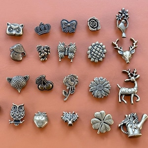 Könnte beinhalten: Eine Sammlung bronzefarbener Metallverzierungen in verschiedenen Formen, darunter Eule, Krone, Schmetterling, Rose, Hirsch und Kleeblatt. Die Verzierungen sind auf einer pfirsichfarbenen Oberfläche angeordnet, die ihre filigranen Designs zeigt.