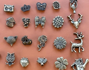 May include: A collection of twenty-four antique bronze-toned metal buttons and charms featuring various shapes including owls, crowns, butterflies, flowers, grapes, cherries, strawberries, deer, a watering can, and circular snap fasteners.