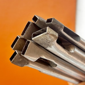May include: A close-up shot of a bundle of metal leathercraft tools. The tools have a rectangular shape with a hollow center and a flat, beveled edge. The tools are made of a silver-colored metal and are arranged against an orange background.