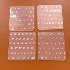 May include: Four clear acrylic letter and number stencils. The stencils include uppercase and lowercase alphabets, numbers, and symbols. The stencils are arranged on a brown surface. The text "Tel:" is visible on one of the stencils.