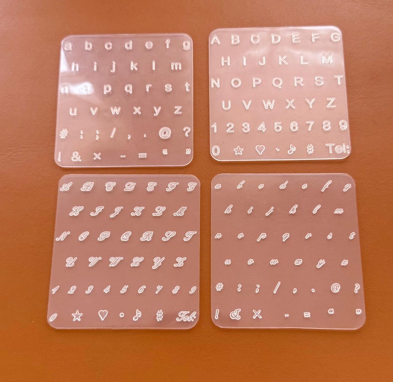 May include: Four clear acrylic letter and number stencils. The stencils include uppercase and lowercase alphabets, numbers, and symbols. The stencils are arranged on a brown surface. The text "Tel:" is visible on one of the stencils.