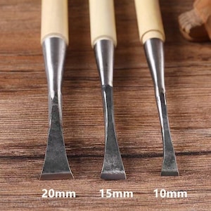 Puede incluir: Primer plano de tres cinceles para tallar madera con hojas de metal y mangos de madera de color claro. Las hojas están etiquetadas con tamaños: 20 mm, 15 mm y 10 mm. Las herramientas están dispuestas sobre una superficie de madera.