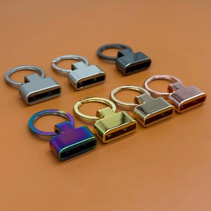 May include: An assortment of metal key fob hardware in various colors, including silver, black, gold, and iridescent. Each piece features a rectangular clasp and a circular key ring, suitable for crafting or personalizing keychains.