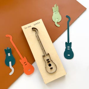 Puede incluir: Un troquel de madera con forma de guitarra, junto con formas de guitarra de cuero recortadas en naranja, verde azulado y verde. El troquel tiene el texto "SX-086 32*153" impreso. El fondo es una mezcla de marrón y blanco.