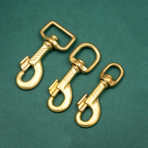 Puede incluir: Tres mosquetones de color latón sobre una superficie verde oscuro. Los ganchos tienen un ojo giratorio y un mosquetón con resorte. El metal tiene un acabado pulido, adecuado para diversas aplicaciones.