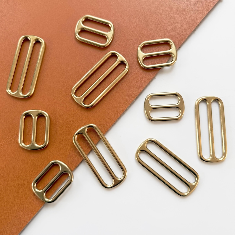 Op de afbeelding: Een verzameling goudkleurige metalen bandglijders in verschillende vormen en maten. De glijders zijn rechthoekig, ovaal en vierkant, met twee of drie sleuven. Ze zijn gerangschikt op een bruin en wit oppervlak.
