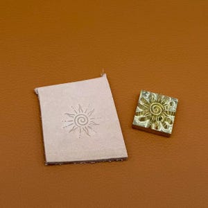 May include: A square brass metal stamp featuring a raised sun design with a central spiral and radiating rays, positioned next to a light beige rectangular piece of leather that has been embossed with the same sun pattern.