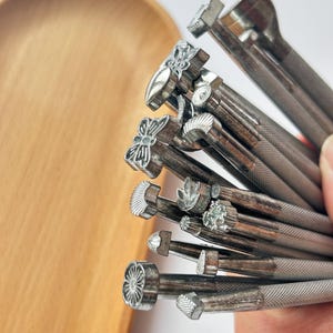 May include: A selection of metal leather stamping tools featuring diverse decorative designs, such as floral and geometric patterns. The tools are held in a hand, with a wooden tray in the background.
