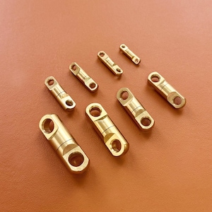 Pode incluir: Uma coleção de conectores giratórios de cor latão, de vários tamanhos, dispostos sobre uma superfície castanha. Cada conector apresenta um corpo cilíndrico com aberturas circulares em cada extremidade, projetadas para fixação. O metal tem um acabamento polido.