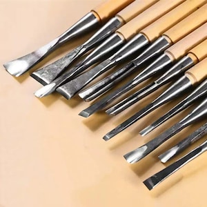 May include: A set of wood carving tools with wooden handles and metal blades. The blades have various shapes, including curved, angled, and flat edges. The tools are arranged diagonally on a light brown surface.