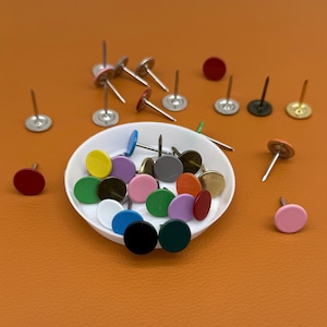 May include: A collection of colorful push pins with round heads in various colors, including red, yellow, green, and pink. Some pins are scattered around a white bowl, while others are arranged on an orange surface.
