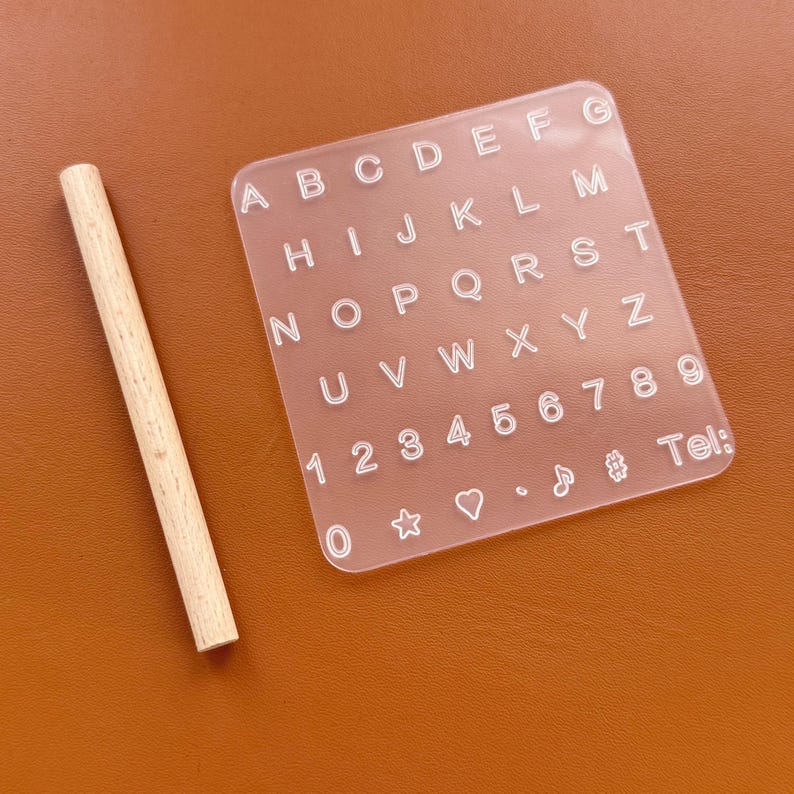 May include: A clear acrylic alphabet and number stencil with a wooden rolling pin. The stencil includes the letters A-Z, numbers 1-9, and symbols. The rolling pin is light brown.