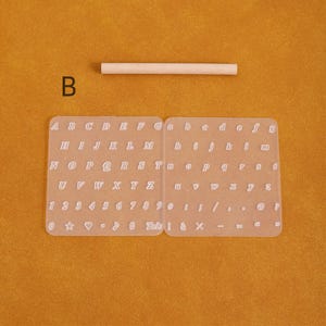 May include: A clear, flexible alphabet and number stencil set with uppercase and lowercase letters, numbers, and symbols. A small wooden dowel is above the stencil. The letter "B" is in the upper left corner.