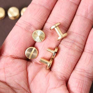 May include: Close-up of several small, gold-colored brass rivets and screws. Some rivets have a slotted head, while others have a decorative sunburst pattern. The screws have a threaded post and a flat head. These fasteners are likely used in leathercraft or other DIY projects.