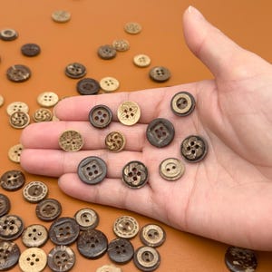 May include: A collection of various brown buttons, including coconut shell and wooden buttons, in different sizes and designs. Some buttons have four holes, while others have decorative patterns. The buttons are scattered on a brown surface.