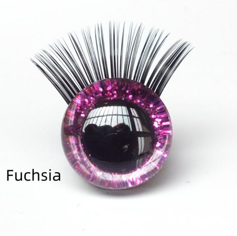 May include: A fuchsia-colored, glitter-infused glass eye with black eyelashes. The eye has a reflective surface and the word "Fuchsia" is printed below it. This item is likely for crafting or doll-making.