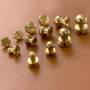 Könnte beinhalten: Eine Sammlung von Messingbeschlägen, darunter Schrauben und dekorative Nieten. Die Hardware hat eine polierte, goldene Oberfläche. Die Teile sind auf einer braunen Oberfläche angeordnet und zeigen ihre verschiedenen Formen und Größen, geeignet für Lederarbeiten oder ähnliche Projekte.