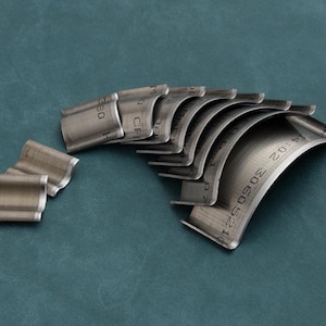 May include: A selection of curved, metallic machine components displayed on a teal background. Each component has a semi-circular form with etched markings, including numbers and letters. The parts are arranged in a stacked, fanned-out manner, highlighting their design and texture.