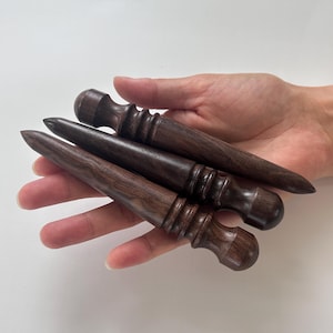 Peut inclure: Quatre outils en bois brun foncé avec des manches arrondis et des pointes effilées sont présentés. Les outils ont une finition lisse et polie et sont tenus dans une main sur fond blanc. Les outils semblent être destinés au travail du cuir ou à des métiers similaires.