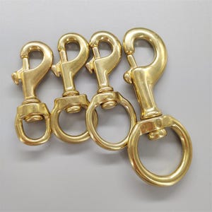 Puede incluir: Cuatro mosquetones de latón de diferentes tamaños están alineados. Cada gancho tiene un cierre de resorte y un ojal giratorio. Los ganchos están hechos de un metal dorado brillante.