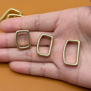 May include: Three gold-colored rectangular metal rings with rounded corners are displayed in a hand. The rings are open in the center and have a smooth, polished finish. The background is a warm, neutral tone.