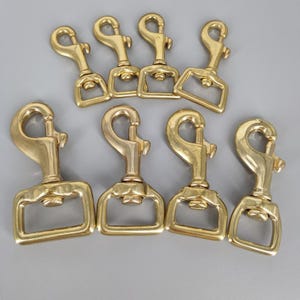 以下が含まれることがあります： 7つの金色のスイベルスナップフック。フックは、一端にスプリング式の留め金、もう一端に長方形のループが付いています。金属製の金具は、灰色の背景に配置されています。