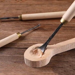 Puede incluir: Primer plano de herramientas para tallar madera con mangos de madera y hojas de metal. Una herramienta está tallando una forma de cuchara de madera, quitando virutas de madera. Las herramientas están sobre una superficie de madera.