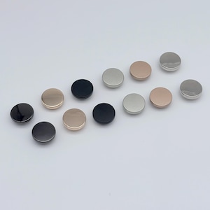 Könnte beinhalten: Eine Sammlung runder Metallknöpfe in verschiedenen Farben, darunter Schwarz, Silber und Roségold. Die Knöpfe sind auf einer weißen Oberfläche angeordnet und zeigen ihre glatten, reflektierenden Oberflächen. Geeignet für Näh- und Bastelprojekte.